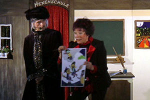 Hexe Schmusemund erklärt den Kindern im Publikum unterschiedliche Hexen und ihre Besen auf einem Plakat. Der Zauberlehrer passt auf.